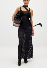 Schwarzes Netz-Top mit einem Choker-Detail, über einem langen schwarzen Spitzenrock geschichtet. Zu den Accessoires gehören eine schwarze Handtasche und silberne Armbänder.