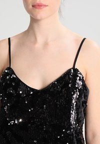 Haut à sequins noirs avec fines bretelles. Présente un décolleté en V, une texture scintillante et une silhouette ajustée. Éléments noirs unis sur les bretelles.