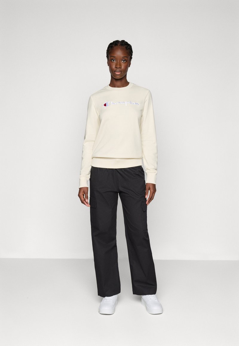 Cremfarbener Sweatshirt mit dem eingestickten "Champion"-Logo in Blau und Rot. Kombiniert mit lockeren, schwarzen Cargo-Hosen. Weiße Sneakers.