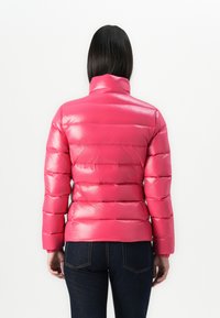Žena s ravnom crnom kosom koja nosi sjajnu ružičastu puffer jaknu i tamne traperice, stoji okrenuta leđima prema posmatraču na bijeloj pozadini.