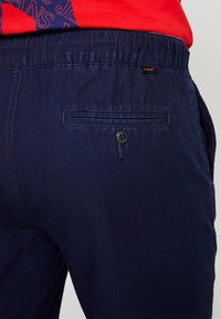 Námořnické modré kalhoty vyrobené z lehkého materiálu s texturovaným povrchem. Obsahují zadní kapsu s knoflíkovým zapínáním a malou značkovou etiketu.
