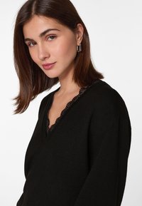 Schwarzer Pullover mit V-Ausschnitt und Spitzenbesatz. Weicher Stoff mit lockerer Passform, langen Ärmeln und einem strukturierten Muster.