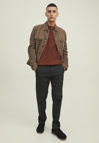 Chaqueta de ante marrón sobre un suéter rojo de canalé, combinado con pantalones negros y zapatos oscuros. El modelo está de pie con las manos en los bolsillos contra un fondo neutro.