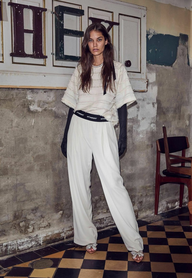 Pantaloni bianchi plissettati con una cintura con logo nero, abbinati a un top strutturato bianco sporco e lunghi guanti neri. Scarpe aperte scintillanti.