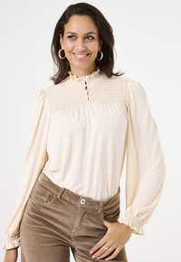 Crème blouse met smock-yoke, gerimpelde kraag en knoopdetails. Lange mouwen met geplooide manchetten. Bruine corduroy broek.