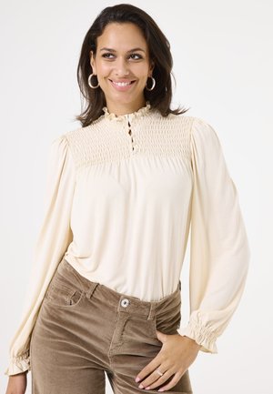 Crème blouse met smock-yoke, gerimpelde kraag en knoopdetails. Lange mouwen met geplooide manchetten. Bruine corduroy broek.