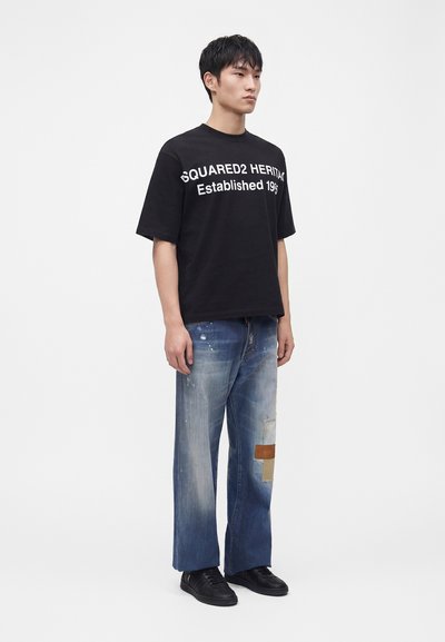 Νέος άνδρας με μαύρο T-shirt με λευκό κείμενο και ξεθωριασμένο μπλε τζιν με πιτσιλιές μπογιάς και μπαλώματα, στέκεται μπροστά σε απλό φόντο.