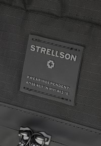 Czarna etykieta z materiału z wytłoczonym napisem "STRELLSON" oraz hashtagiem "#WEARINDEPENDENT", z współrzędnymi geograficznymi i małym emblematem.