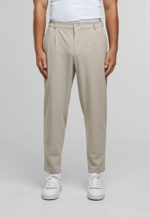 Pantalones beiges ajustados con pliegues, cinturilla elástica y botón delantero. Llevados con zapatillas blancas, con una textura suave.