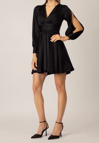 Svart satinklänning med V-ringning, figurnära midja och utställd kjol. Långa puffärmar har utskärningar. Matchas med svarta klackskor med spetsig tå.