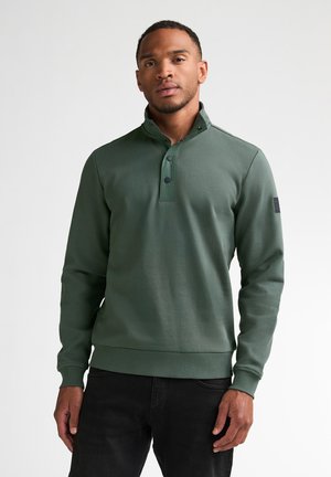 Man draagt een groene trui met lange mouwen, een hoge kraag en drie zwarte knopen, gecombineerd met een zwarte broek, staand tegen een witte achtergrond.