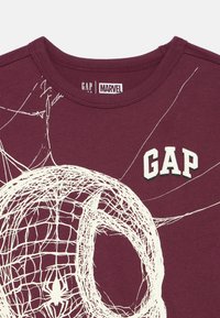 Bordové tričko s okrúhlym výstrihom zdobené bielou grafikou pavúčej siete a nápisom "GAP" v štýlovom písme. Vyrobené z mäkkej bavlny.