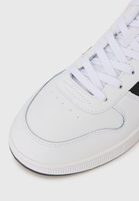 White sneaker featuring a smooth leather upper, round toe, and white laces. Notable black accent near the heel and textured rubber sole.