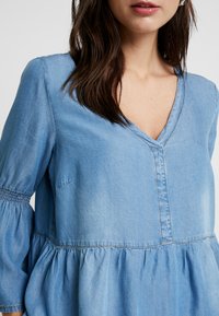 Light blue denim top with a V-neck, smocked sleeves, and a gathered peplum hem. Features a front seam and soft texture.