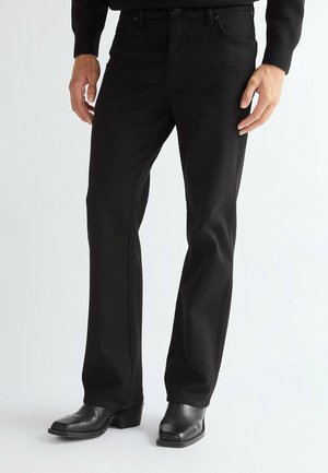 Uomo che indossa jeans neri a gamba dritta e stivali neri in pelle con punta quadrata su uno sfondo chiaro e semplice.