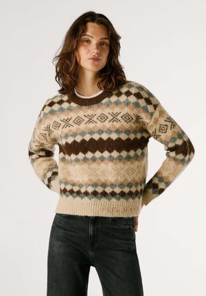 Pull en tricot beige et brun avec des motifs en diamants et géométriques. Équipé d'un col rond et de poignets côtelés. Porté par-dessus un jean noir.