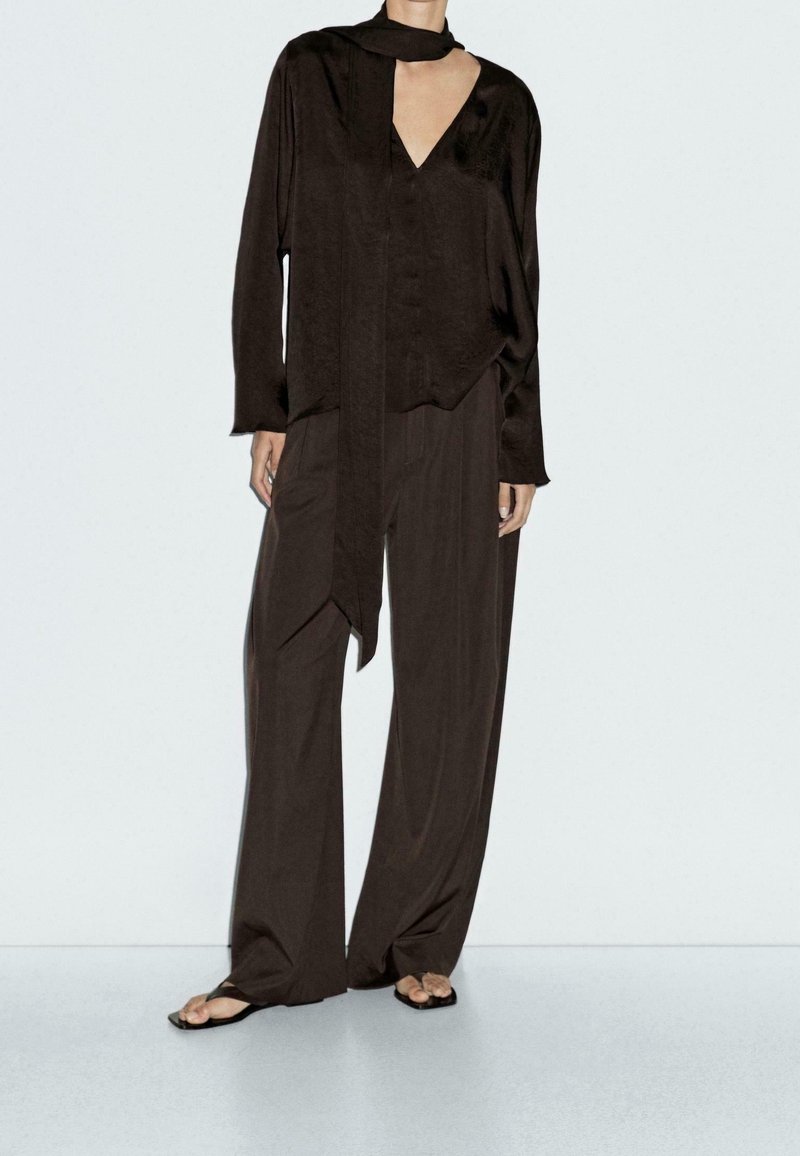 Personne portant un chemisier noir ample à col en V avec un long détail d’écharpe et un pantalon noir à jambes larges avec des sandales noires à bout ouvert.