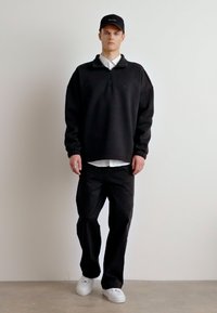Felpa nera con zip e colletto, abbinata a pantaloni neri e sneakers bianche. Il modello indossa un cappellino nero, posato su uno sfondo neutro.