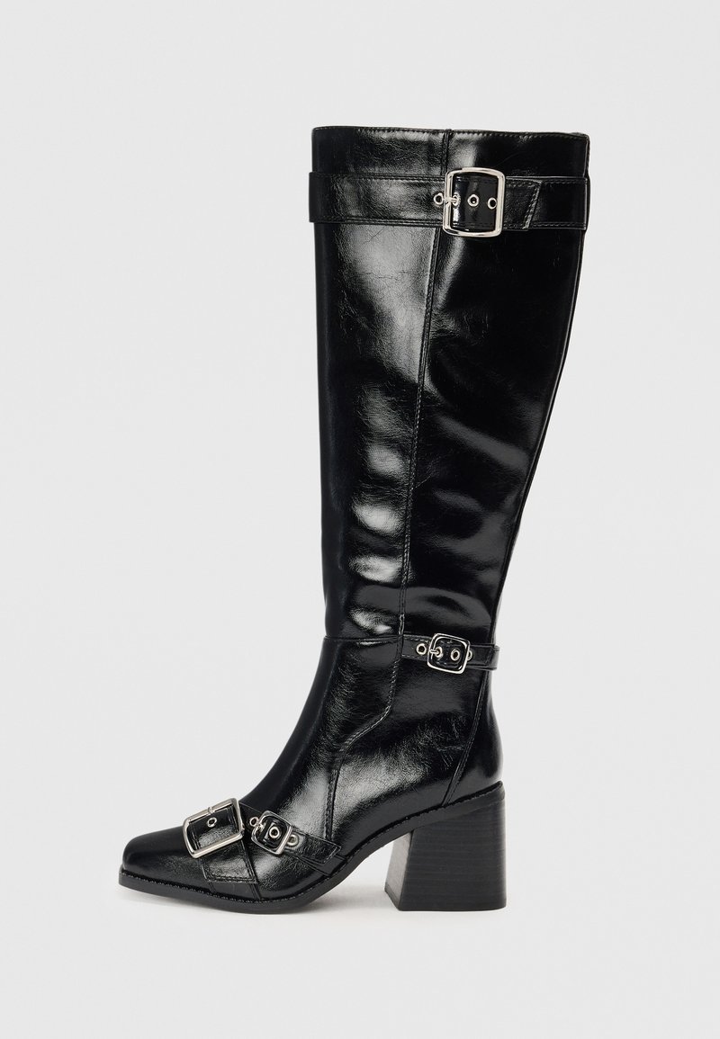 Bota negra de cuero hasta la rodilla con un acabado brillante. Presenta hebillas y correas plateadas, una puntera afilada y un tacón grueso y texturizado.