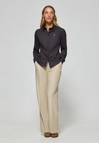 Chemise noire à boutons, en tissu lisse, avec un col et un détail de logo blanc, assortie à un pantalon large beige et des chaussures marron.