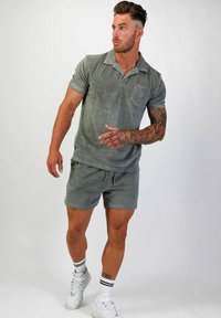 Grå frotté polo med bröstficka, kombinerad med matchande shorts. Har en avslappnad passform och sidodragband. Vita sneakers bärs.