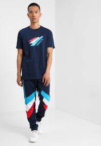 Maglietta in cotone blu navy con logo adidas multicolore, abbinata a pantaloni da jogging con audaci motivi geometrici in rosso, blu e bianco.