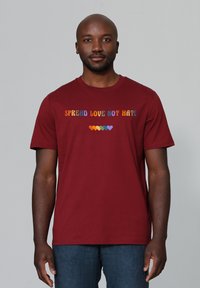 Bordeaux katoenen T-shirt met meerkleurige tekst "SPREAD LOVE NOT HATE" boven een rij kleurrijke harten. Korte mouwen en ronde halslijn.