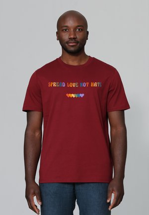 T-shirt en coton bordeaux avec le texte multicolore "RÉPANDRE L'AMOUR PAS LA HAINE" au-dessus d'une rangée de cœurs colorés. Manches courtes et col rond.