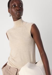 Bež majica brez rokavov z visokim ovratnikom, z gladko teksturo in subtilnimi tonami. V kombinaciji z svetlo obarvano torbico.