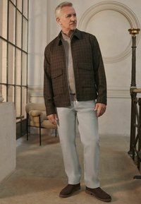 Brown plaid zip-up jacket over gray sweater, light gray trousers, and dark suede shoes. Setting features neutral tones and modern architecture.
