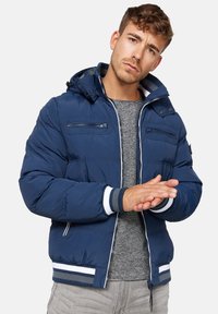 Mørkeblå puffjakke med lynlås foran, justerbar hætte, stribede detaljer og ribbede manchetter, båret over en grå sweater.