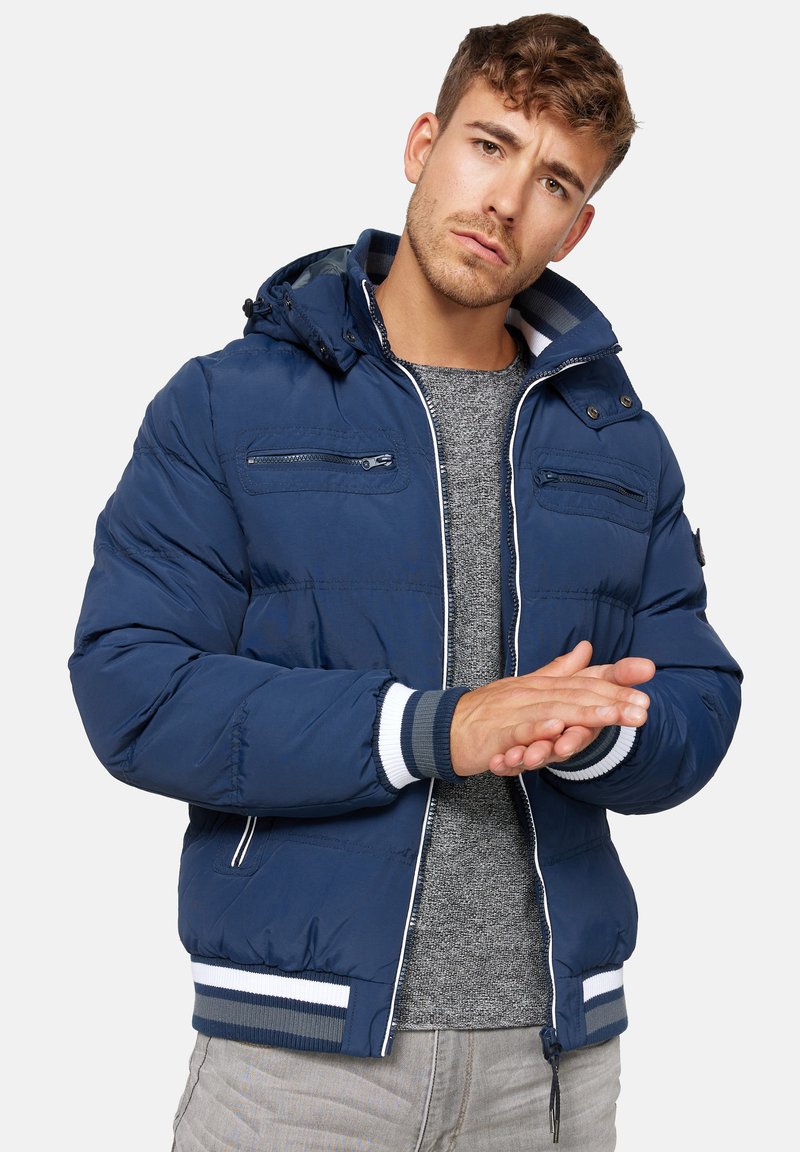 Mørkeblå puffjakke med lynlås foran, justerbar hætte, stribede detaljer og ribbede manchetter, båret over en grå sweater.