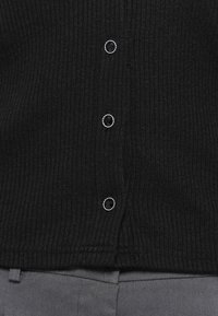 Tissu côtelé noir avec trois boutons ronds noirs bordés d'argent, au-dessus d’un pantalon en tissu gris.