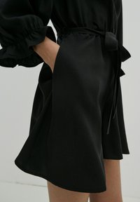 Zwarte jurk met een getailleerde taille met een riem, uitlopende rok en gerimpelde mouwmanchetten. Gladde textuur met minimale details.