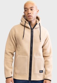 Beżowy polarowy hoodie z czarnym zamkiem z przodu, kapturem na sznurki i dwiema dużymi kieszeniami z przodu. Teksturowany materiał z czarnymi akcentami.