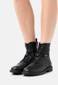 Botas de tobillo de cuero negro con cordones y una cremallera lateral. Presentan un acabado texturizado, suela de goma y detalles en tono dorado.