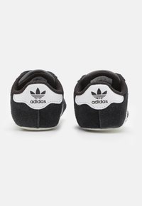 Černé semišové tenisky s bílým akcentem na patě a ikonickým trojlístkovým logem. Měkká textura s polstrovaným okrajem pro pohodlí.