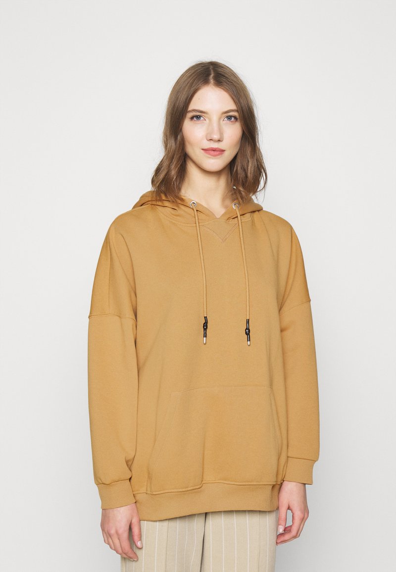 Hoodie boje senfa s V-izrezom, vezicama i prednjim džepom. Mekani, pamučno-sintetički materijal i oversized kroj s dugim rukavima.