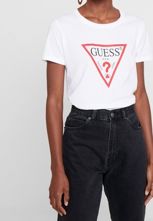 Femme portant un t-shirt blanc avec le logo Guess rentré dans un jean noir taille haute, visible des épaules aux hanches sur un fond uni.