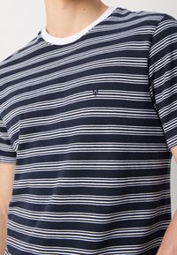 T-shirt à rayures horizontales bleu marine et blanches avec col blanc porté par une personne, avec un petit logo brodé sur la poitrine.