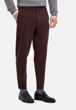 Mann, der braune maßgeschneiderte Hosen, graue Socken, schwarze Schlupfschuhe und einen grauen Pullover trägt und mit einer Hand in der Tasche steht.