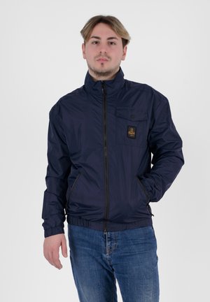 Giacca leggera blu navy con colletto alto, chiusura con zip, tasca frontale con bottone a pressione e orlo elasticizzato. Indossata sopra dei jeans in denim.