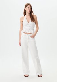Top halter bianco con peplum e motivo ad occhio, abbinato a jeans bianchi larghi. Sandali con cinturini e orecchini floreali completano il look.