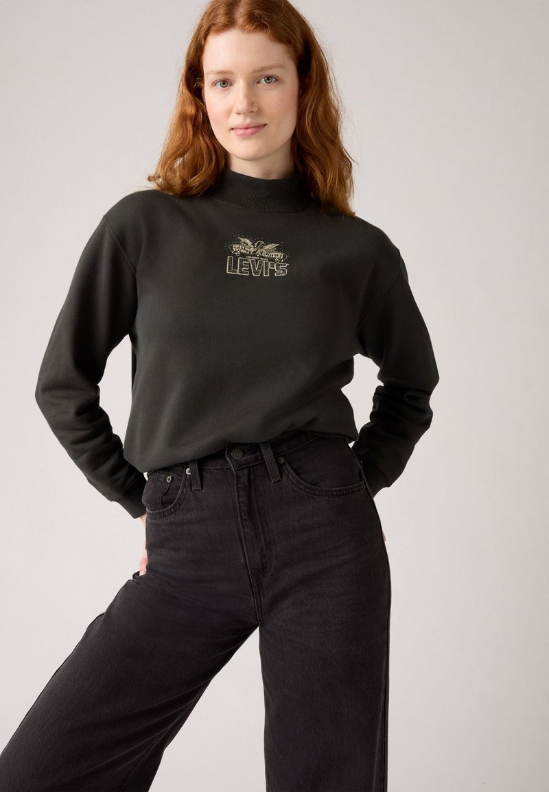 Fekete pamut pulóver magas nyakkal és arany "Levi's" logóval, magas derekú fekete farmernadrággal párosítva.