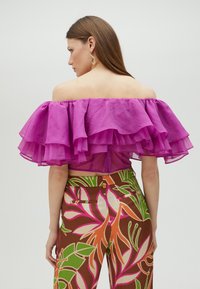 Skulderfri bluse i gennemsigtig, lys pink stof med lagdelte flæser; parret med trykte brune bukser med store blomsterdesigns i forskellige farver.