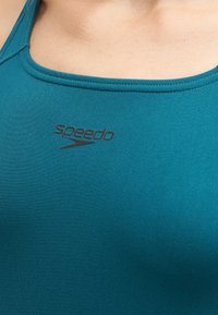 Κοντινή λήψη του υφάσματος μαγιό Speedo σε τιρκουάζ απόχρωση που καλύπτει το στήθος και τους ώμους ενός ατόμου με ορατές ραφές στο ντεκολτέ.