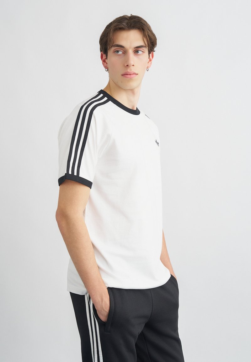 Biela tričko s čiernym lemovaním a tromi čiernymi pruhmi pozdĺž ramien. Vyrobené z hladkej látky s priliehavým strihom.
