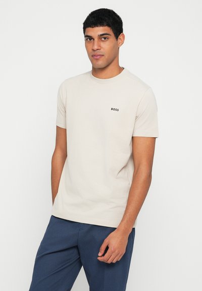 T-shirt beige à manches courtes en coton, avec un col rond et un petit logo noir "BOSS" sur la poitrine. Texture lisse.