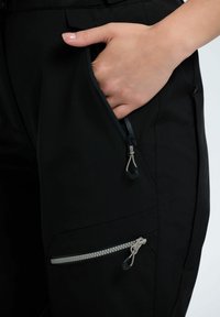 Hand mit manikürten Nägeln teilweise im offenen Reißverschlussfach einer schwarzen Hose mit zwei grauen Reißverschlusszügen.