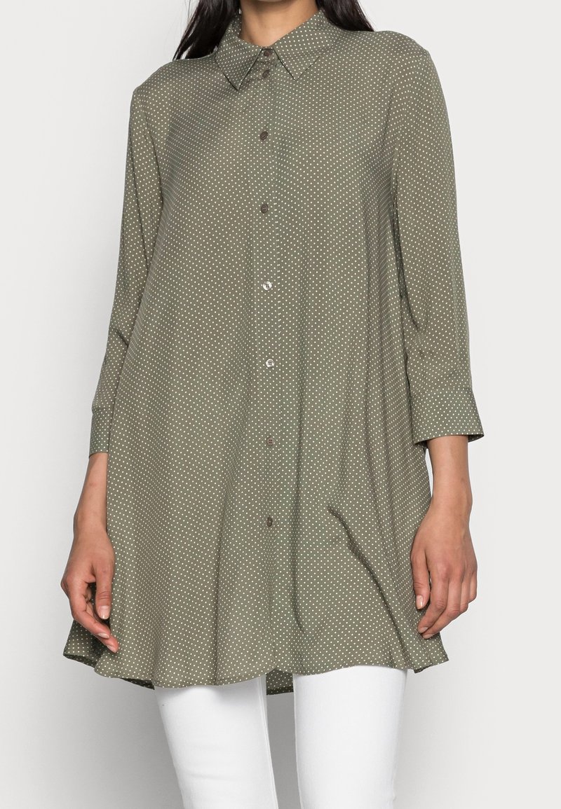 Olive green button-up shirt with a collar, three-quarter sleeves, and white polka dots. Soft fabric with a slightly flared hem.